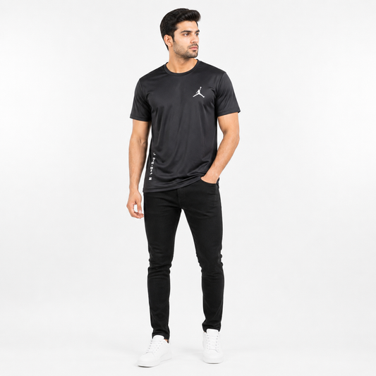 A man modeling a black short-sleeve athletic t-shirt with a white Jumpman logo on the chest, paired with black pants and white sneakers, against a plain white studio background.