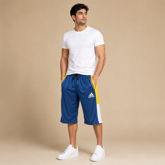 A man modeling blue Adidas athletic shorts with yellow and white side panels, paired with a white t-shirt and white sneakers, standing against a solid beige studio background.