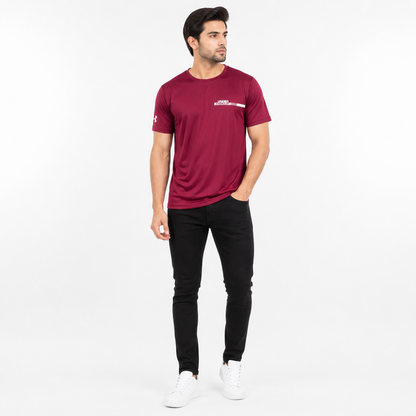 A man modeling a maroon athletic t-shirt with a white Under Armour logo on the chest and sleeve, paired with black pants and white sneakers, against a plain white studio background.