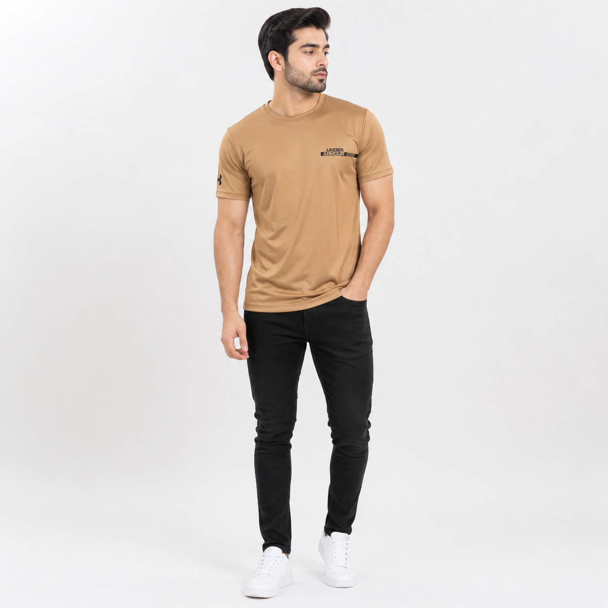 A man modeling a tan crew-neck t-shirt with a small black graphic on the left chest, paired with black pants and white sneakers, against a plain light gray background.