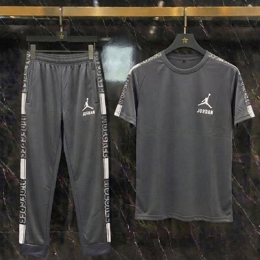 A dark gray two-piece athletic set featuring a short-sleeve t-shirt and matching sweatpants, both hanging on black hangers against a dark marble wall. The t-shirt and pants have white Jordan Jumpman logos and 'JORDAN' text on the chest and sleeve cuffs.