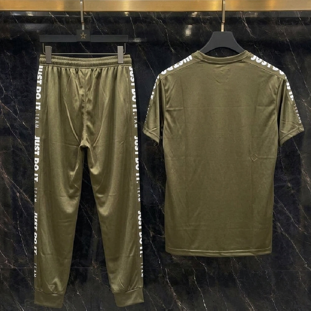 An olive green two-piece tracksuit set consisting of a short-sleeve t-shirt and matching jogger pants, displayed on black hangers against a dark marble-patterned wall. The jogger pants feature white text down the sides, and the t-shirt has white text printed along the shoulders.