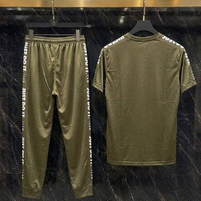An olive green two-piece tracksuit set consisting of a short-sleeve t-shirt and matching jogger pants, displayed on black hangers against a dark marble-patterned wall. The jogger pants feature white text down the sides, and the t-shirt has white text printed along the shoulders.