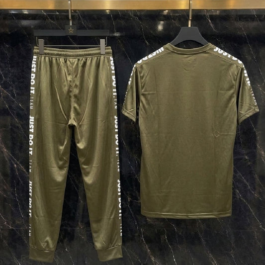 An olive green two-piece tracksuit set consisting of a short-sleeve t-shirt and matching jogger pants, displayed on black hangers against a dark marble-patterned wall. The jogger pants feature white text down the sides, and the t-shirt has white text printed along the shoulders.