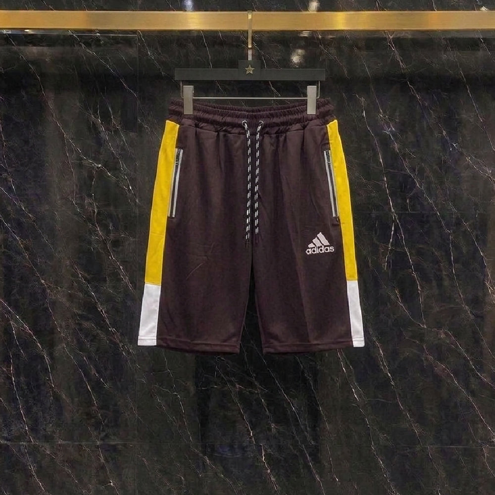 A pair of Adidas athletic shorts in dark brown, featuring yellow and white side panels, hanging on a black hanger against a dark marble-patterned wall.