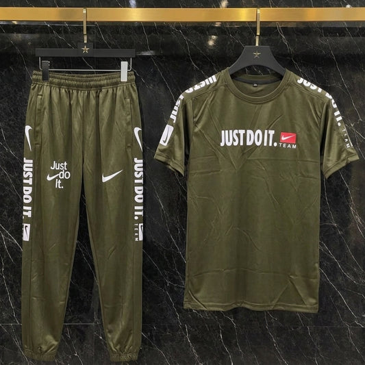 An olive green Nike tracksuit set consisting of a short-sleeve t-shirt and matching jogger pants, displayed on hangers against a black marble background. The t-shirt features 'JUST DO IT.
