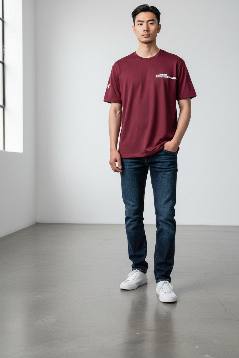 A man wearing a burgundy short-sleeve t-shirt with a small white chest logo, dark blue jeans, and white sneakers, standing in a minimalist studio with a gray floor and white walls.