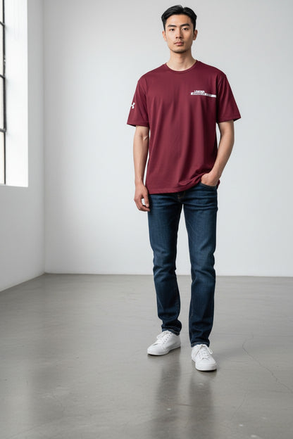A man wearing a burgundy short-sleeve t-shirt with a small white chest logo, dark blue jeans, and white sneakers, standing in a minimalist studio with a gray floor and white walls.