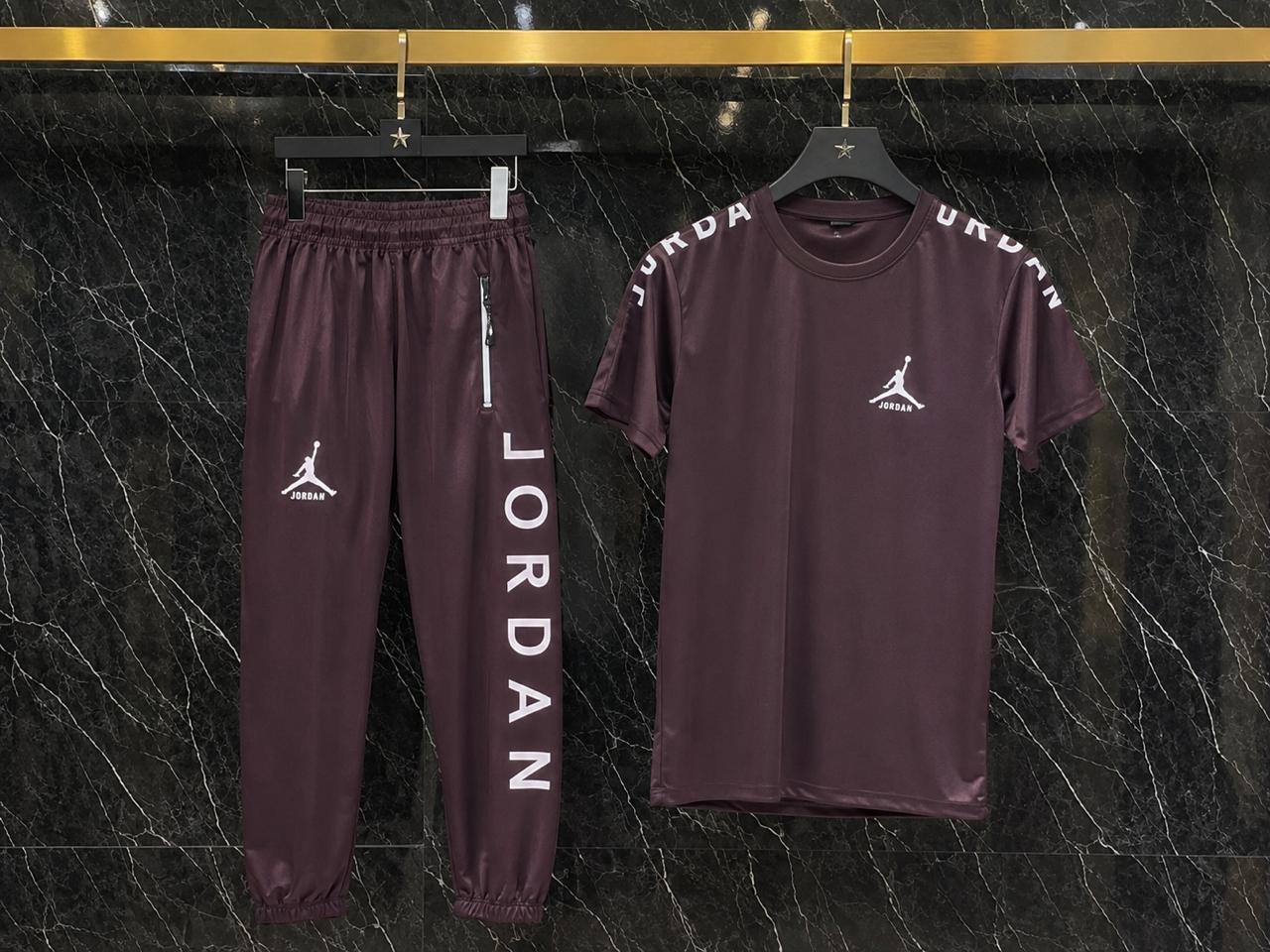 A matching two-piece athletic tracksuit set by Jordan, featuring a short-sleeve t-shirt and jogger pants in a deep plum purple color. The t-shirt has 'JURDAN' printed across the shoulders and a Jumpman logo on the chest, while the pants feature 'JORDAN' printed vertically on one leg and a Jumpman logo on the other leg.