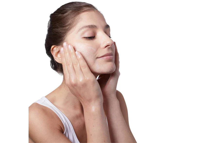 A woman with her eyes closed applies a glossy, dewy skincare product to her face with her hands, set against a solid white background.